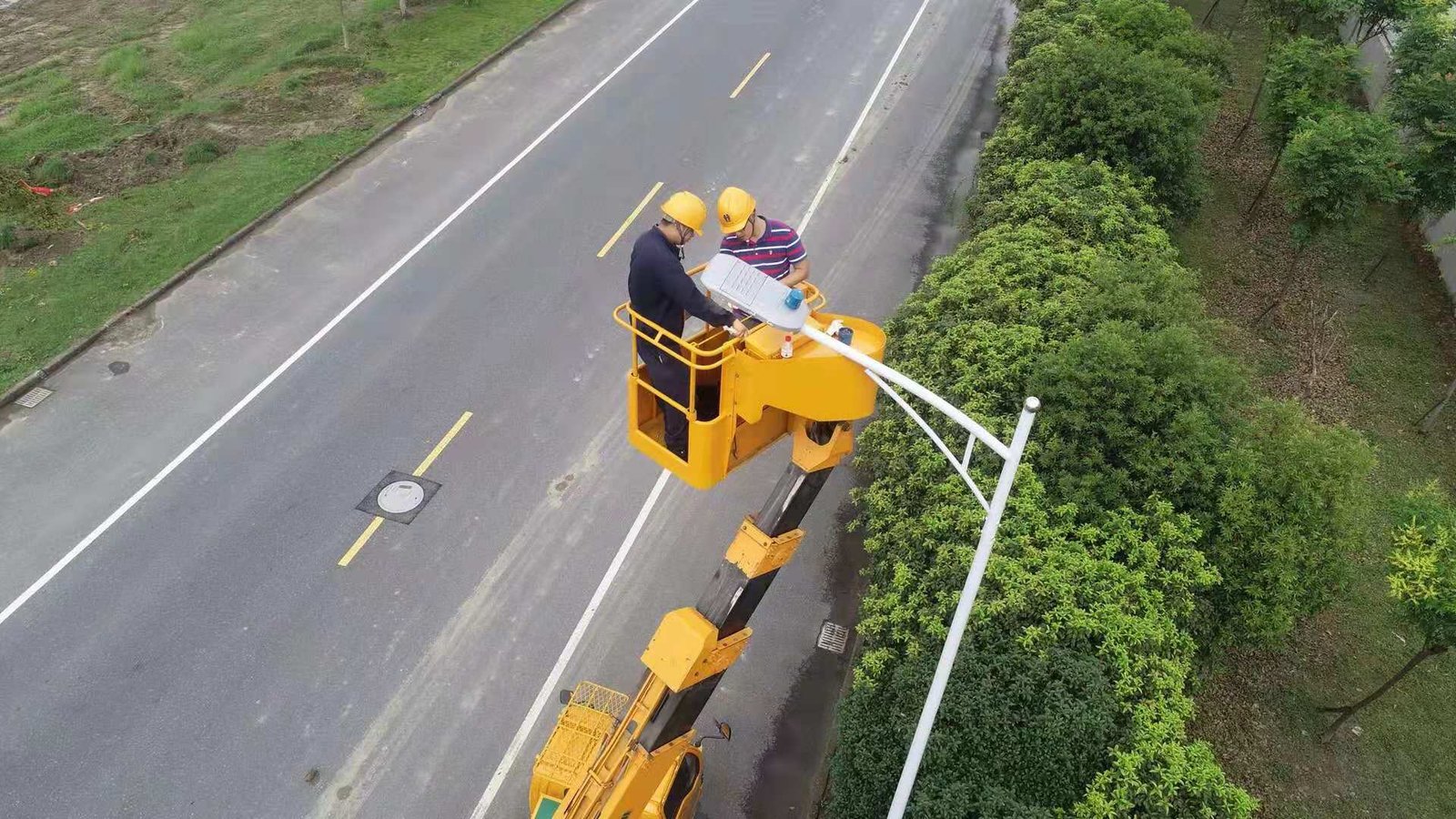 Workers beside a road luminaire on an elevated work platform, with a blue control device visible above the lamp.